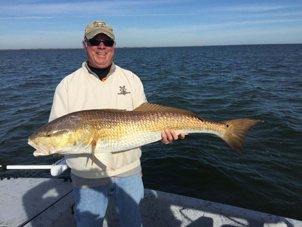 A smiling fisherman in a cap and sunglasses, standing on a boat, holding a Redfish