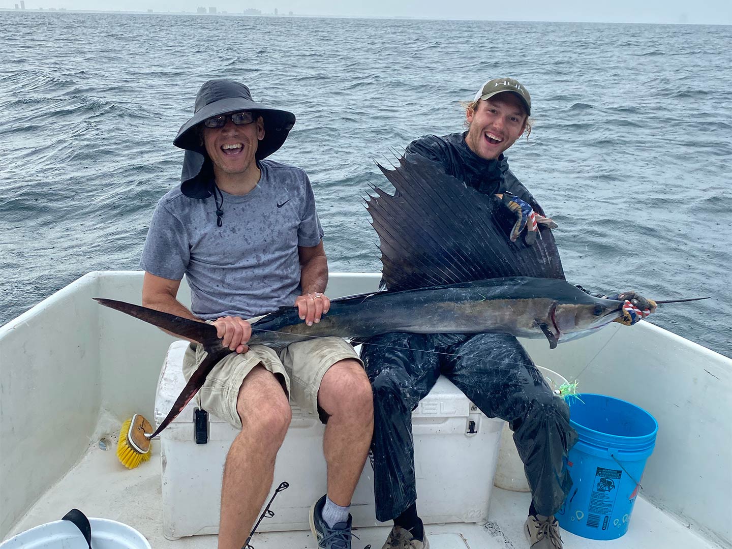 Two anglers hold a Sailfish aboard a boat with Gulf Shores in the distant background