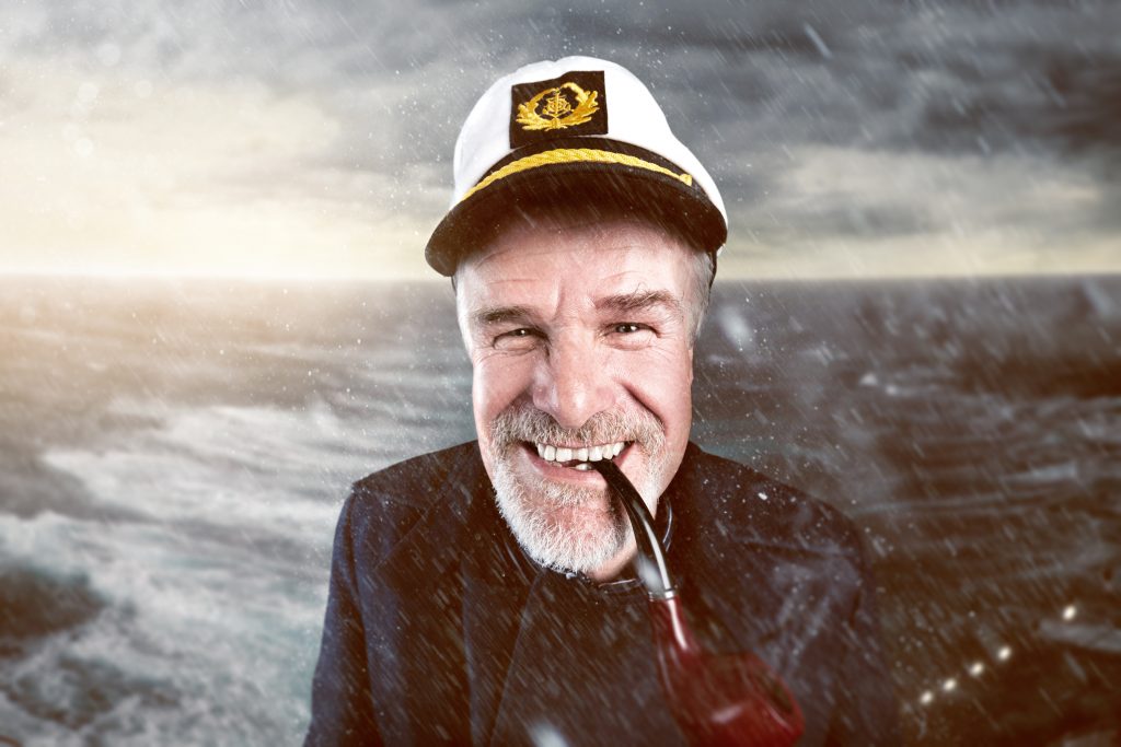 A grinning sea captain with a pipe in his mouth standing on the deck of a ship in a storm