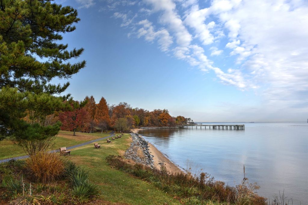 A view of the Chesapeake Bay shoreline, with benches overlooking a small beach and a small fishing pier sticking out in the distance