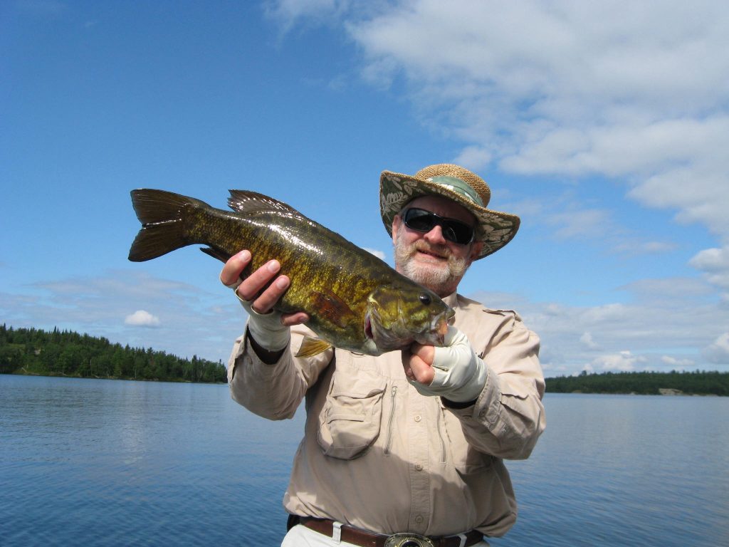 A smiling fisherman holding a Smallmouth Bass with blue skies in the background