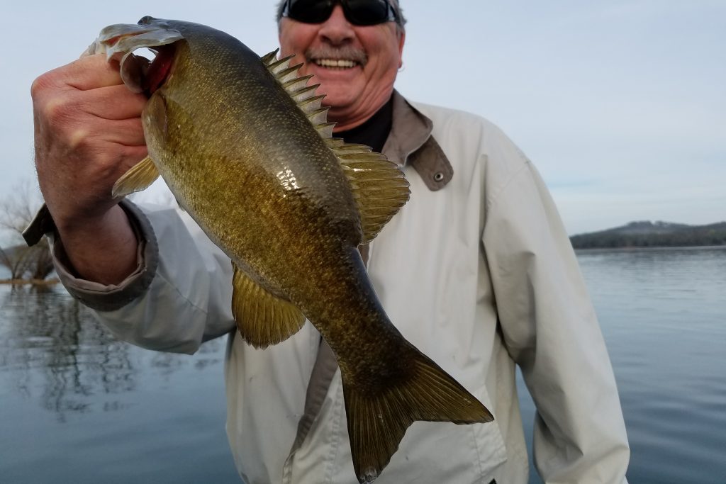 A Smallmouth Bass being held up by a happy angler in a white jacket, with water behind him