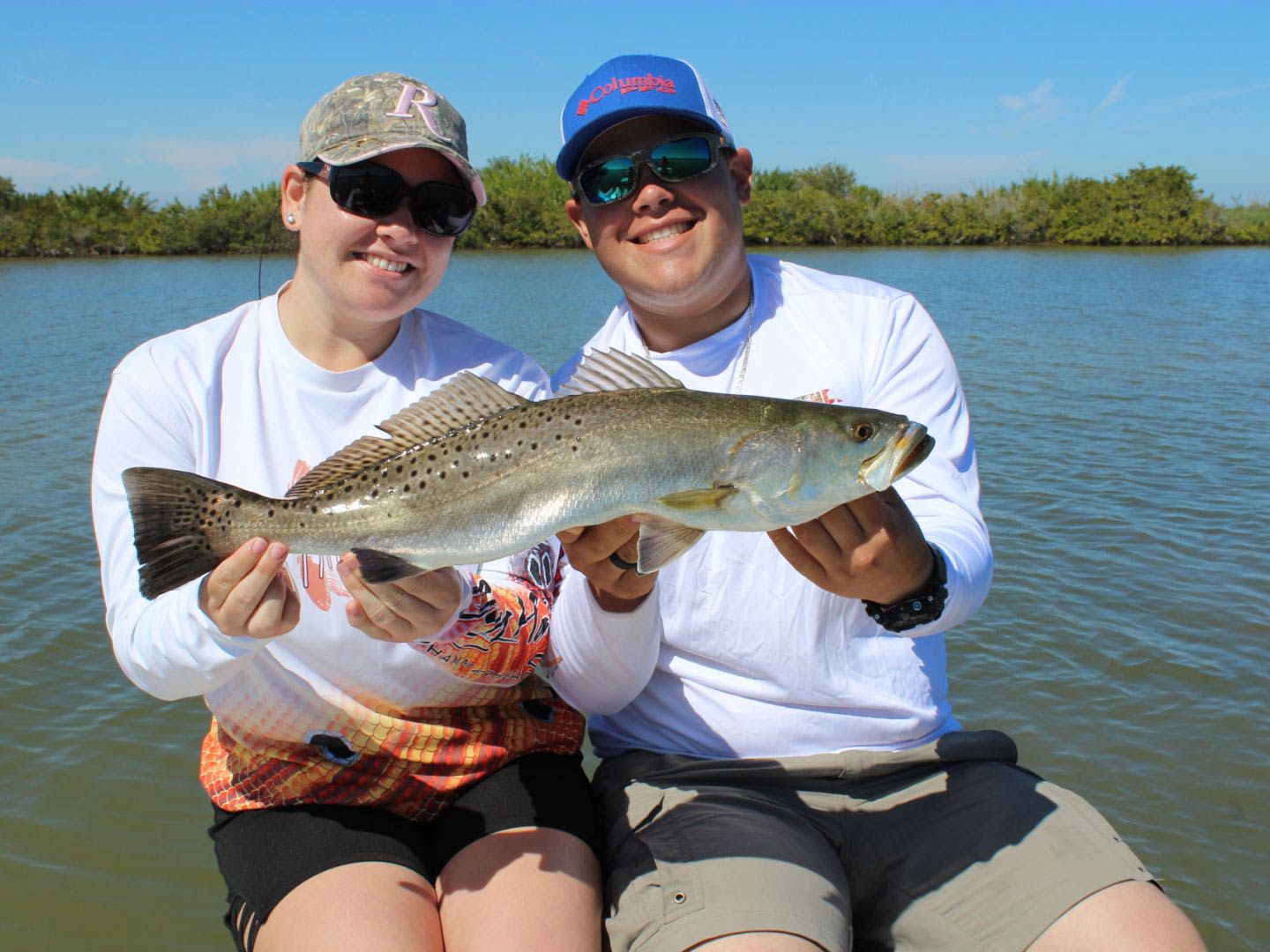 Ein lächelndes Paar sitzt an Bord eines Bootes und hält eine Speckled Trout mit dem Indian River im Hintergrund