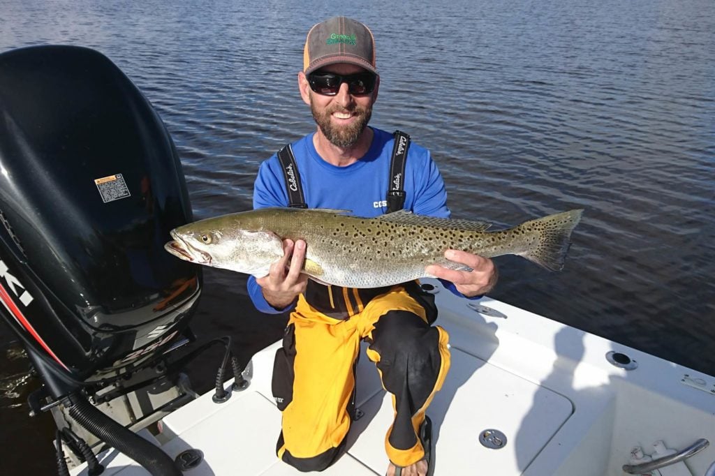 A smiling fisherman holding a big Spotted Seatrout while standing in the corner of a boat