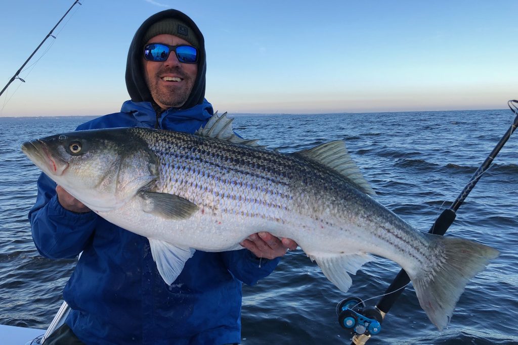 A smiling angler ona  boat wearing a winter jacket, sunglasses, and a hood, holding a trophy Striped Bass with the ocean behind him