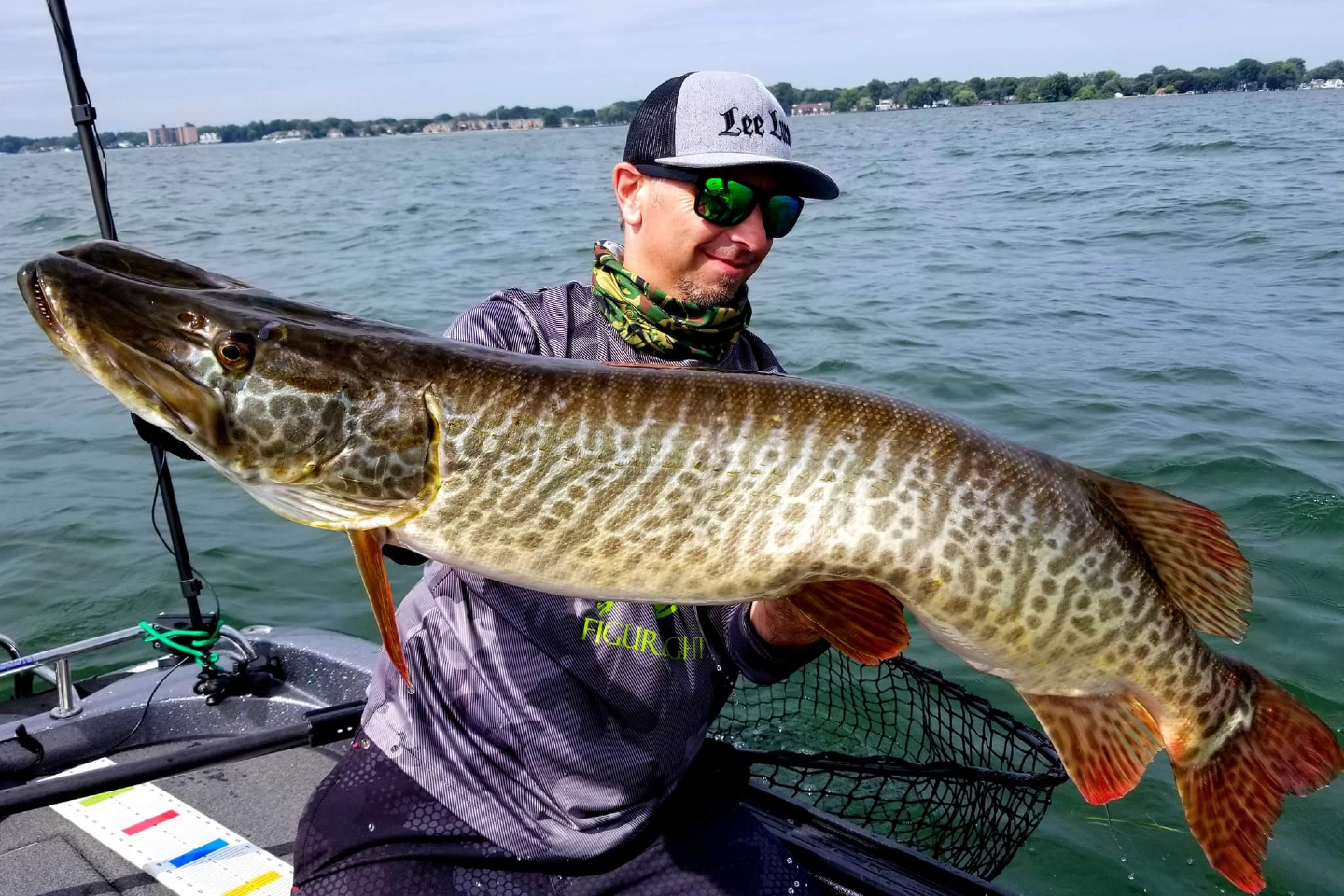 A fisherman in a cap and shades holding a large Tiger Muskie fish with water in the background.