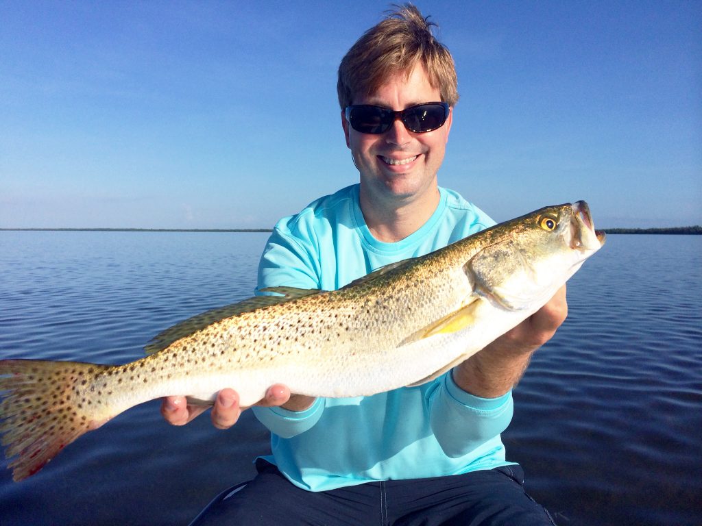 A satisfied angler holding a big Spotted Seatrout with calm waters and blue skies in the background