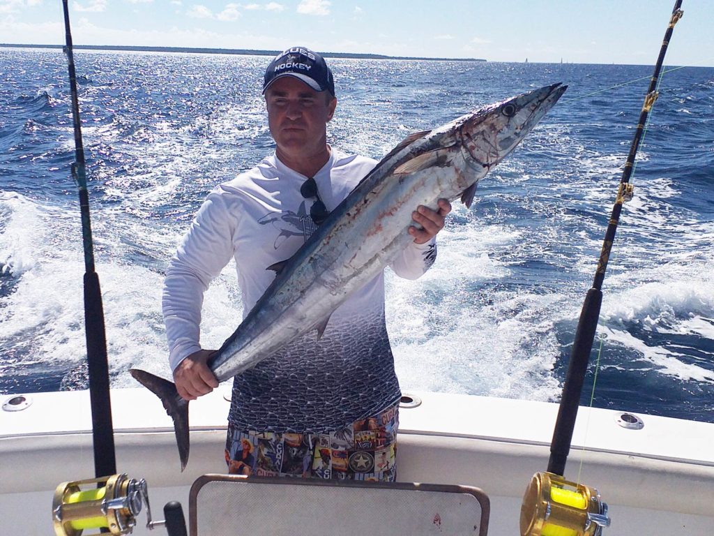 Un pescador sosteniendo, parado en un bote, entre dos cañas, sosteniendo un Wahoo