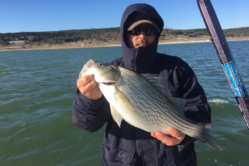 An angler in a black jacket holding a White Bass with water behind him