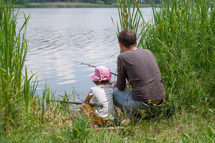 A dad and his daughter fishing together from a bank of a lake.