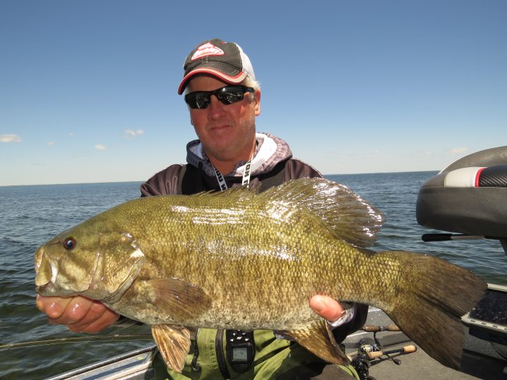 An angler wearing sunglasses and a cap holding a smallmouth bass with open water behind him