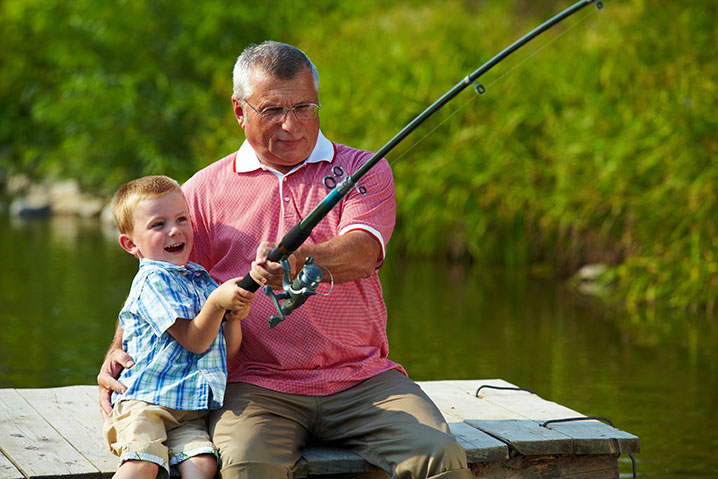 A grandfather teaching his grandson how to fish on a river bank