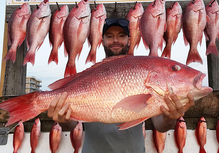 Pescador sosteniendo un pargo rojo con un estante detrás de él lleno de otros pargos, todos capturados a bordo de un barco de fiesta en Destin, Florida.