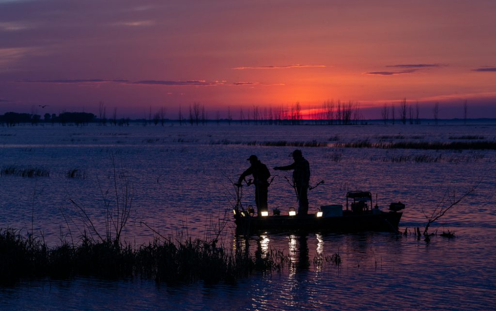 two bowfishers on a small boat, fishing with the sunset in the background