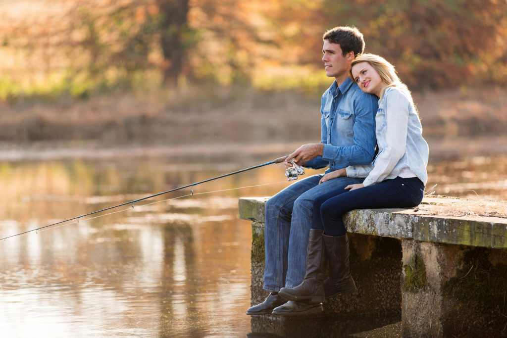 A couple sitting on a dock while the man holds a fishing rod.