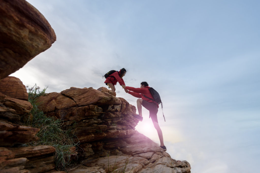 A girl hiker is standing on a top stone and helping out another hiker climb up.