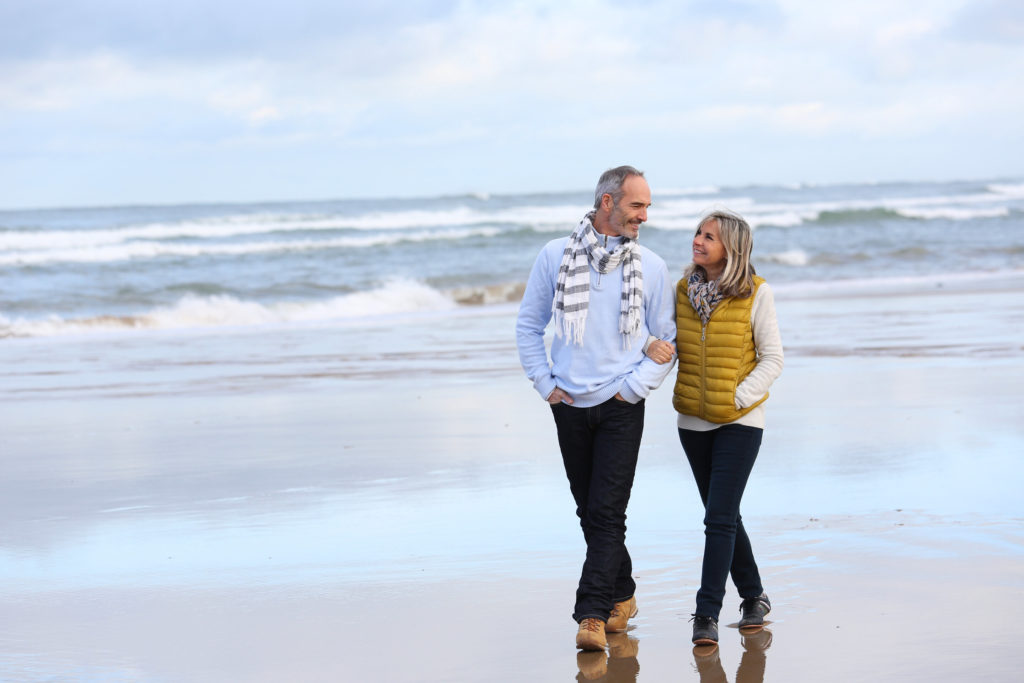 A senior couple walking together on a beach during a Valentine's Day date with waves behind them. 
