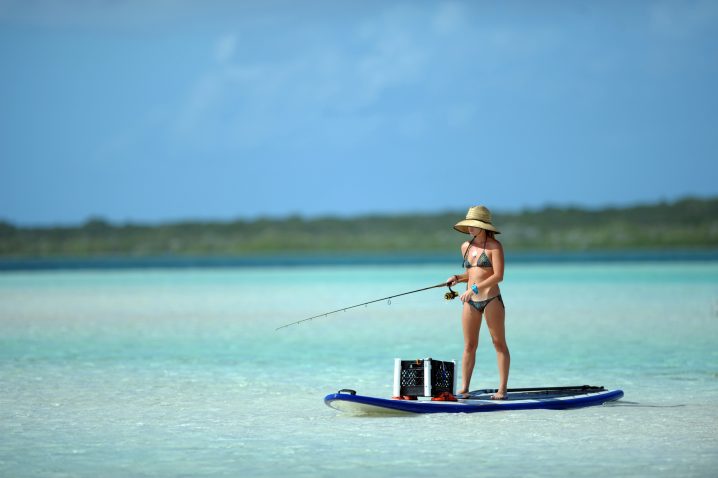 A woman in a bikini and a hat holding a fishing rod on a stand up paddle board