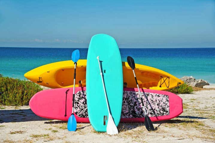 A turquoise stand up paddle board leaning on a pink SUP board and a yellow kayak with the sea in the background