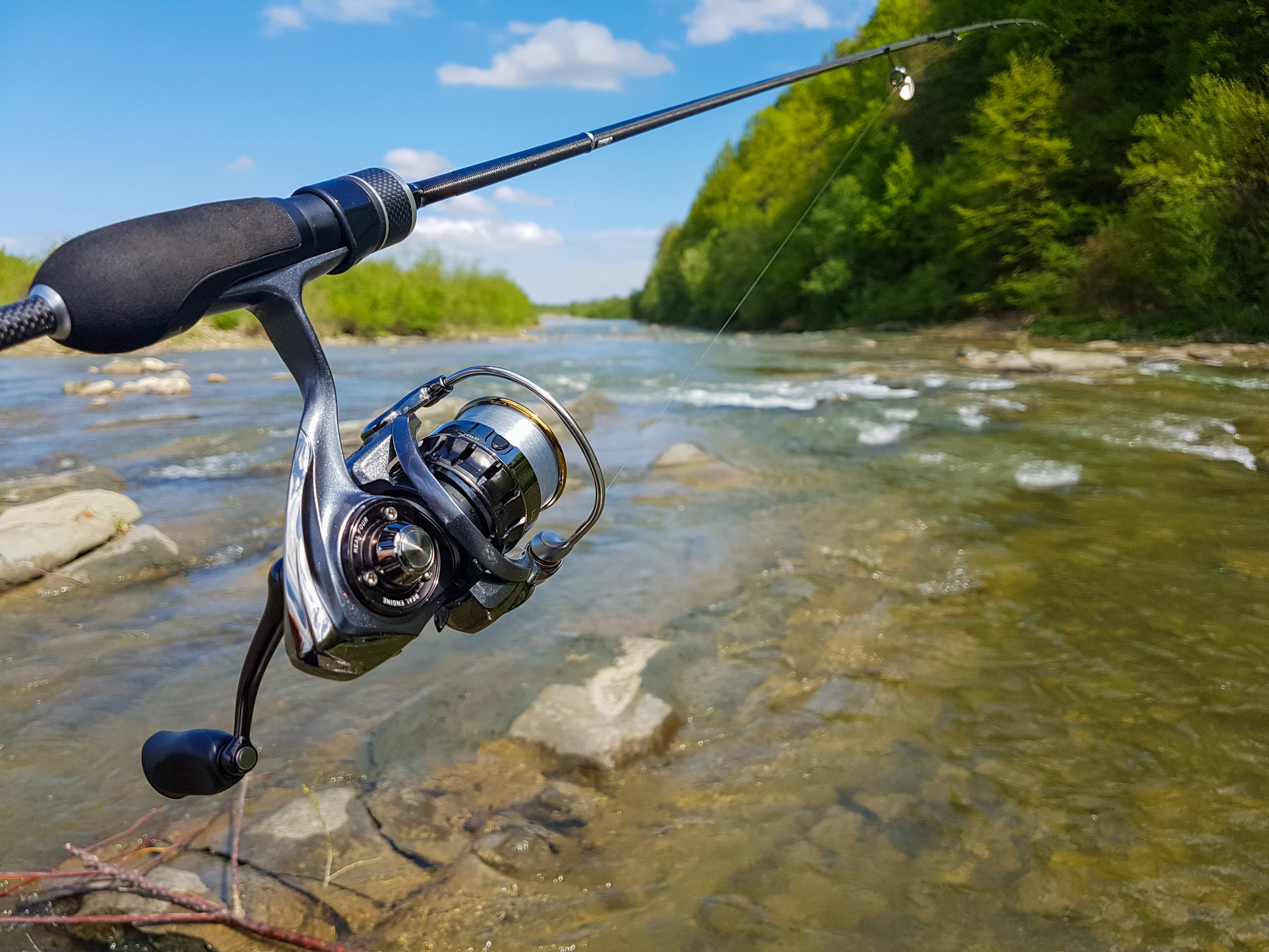 a closeup of a spinning reel on a fishing rod with a river in the background