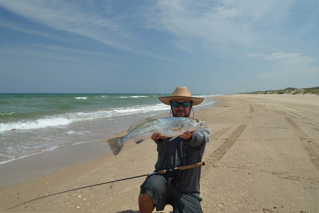A man in a hat and sunglasses holding a Speckled Trout caught while fly fishing on a beach in Texas